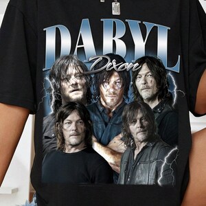 Puede incluir: Camiseta negra con un gráfico de Daryl Dixon de The Walking Dead. El diseño incluye el nombre "DARYL" en azul y plata, con "Dixon" debajo. La camiseta tiene un aspecto vintage y desgastado.