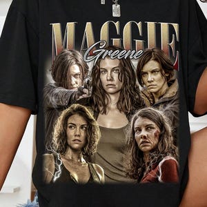 Puede incluir: Camiseta negra con un gráfico del personaje Maggie Greene de The Walking Dead. El diseño incluye el nombre "MAGGIE Greene" en letras doradas sobre un collage de imágenes del personaje. La camiseta tiene mangas cortas.