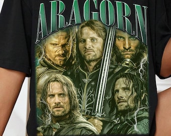 May include: Black t-shirt featuring the word "ARAGORN" in green lettering above a graphic of several characters. The graphic is a movie poster style image. The shirt has short sleeves and a crew neck.