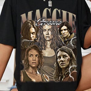 Puede incluir: Camiseta negra con un gráfico del personaje Maggie Greene de The Walking Dead. El diseño incluye múltiples imágenes del personaje con el nombre "Maggie Greene" en una fuente de estilo vintage. La camiseta tiene mangas cortas.