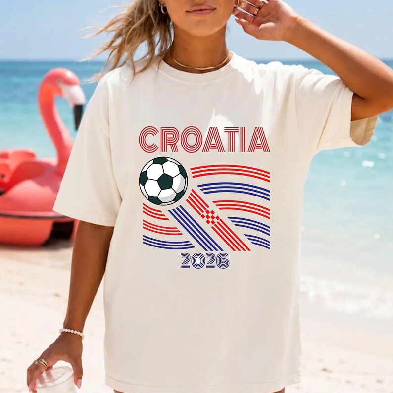 May include: An off-white short-sleeve t-shirt featuring the text CROATIA in red, a black and white soccer ball, and a graphic of red and blue horizontal lines with the year 2026 printed in blue at the bottom center of the design.