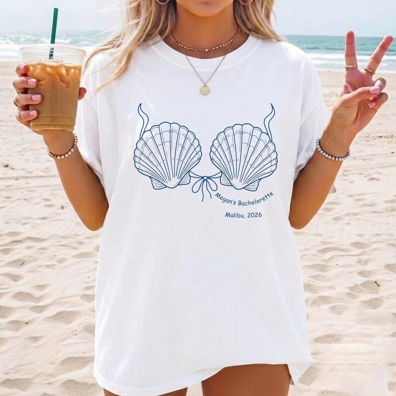 May include: A white t-shirt featuring a blue graphic of two seashells arranged like a bikini top. Below the shells, the text reads Megan's Bachelorette and Malibu, 2026. The shirt has a crew neckline and short sleeves, presented in a casual style.