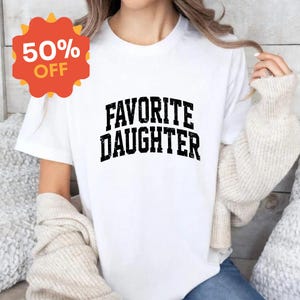 Puede incluir: Camiseta blanca con la frase "FAVORITE DAUGHTER" en letras negras y gruesas con efecto desgastado. Una pegatina roja y naranja con el 50% de descuento est&aacute; en la esquina superior izquierda. La camiseta est&aacute; siendo usada.