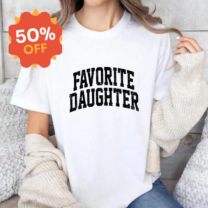 Puede incluir: Camiseta blanca con la frase "FAVORITE DAUGHTER" en letras negras y gruesas con efecto desgastado. Una pegatina roja y naranja con el 50% de descuento est&aacute; en la esquina superior izquierda. La camiseta est&aacute; siendo usada.