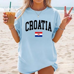 K&ouml;nnte beinhalten: Ein hellblaues Kurzarm-T-Shirt mit dem Wort CROATIA in gro&szlig;en schwarzen Gro&szlig;buchstaben auf der Brust, platziert &uuml;ber einer kleinen, rechteckigen Grafik der kroatischen Nationalflagge mit roten, wei&szlig;en und blauen Streifen.