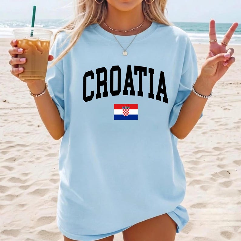 K&ouml;nnte beinhalten: Ein hellblaues Kurzarm-T-Shirt mit dem Wort CROATIA in gro&szlig;en schwarzen Gro&szlig;buchstaben auf der Brust, platziert &uuml;ber einer kleinen, rechteckigen Grafik der kroatischen Nationalflagge mit roten, wei&szlig;en und blauen Streifen.