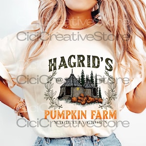 May include: Cream-colored t-shirt featuring a graphic design. The design includes the text "Hagrid's Pumpkin Farm" with an illustration of a cottage, pumpkins, and trees. The text "Magically Grown" is also included.