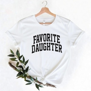 Puede incluir: Una camiseta blanca con el texto "FAVORITE DAUGHTER" en una fuente negra desgastada. La camiseta est&aacute; colgada de una percha de madera. Hay vegetaci&oacute;n en la esquina inferior izquierda de la imagen.
