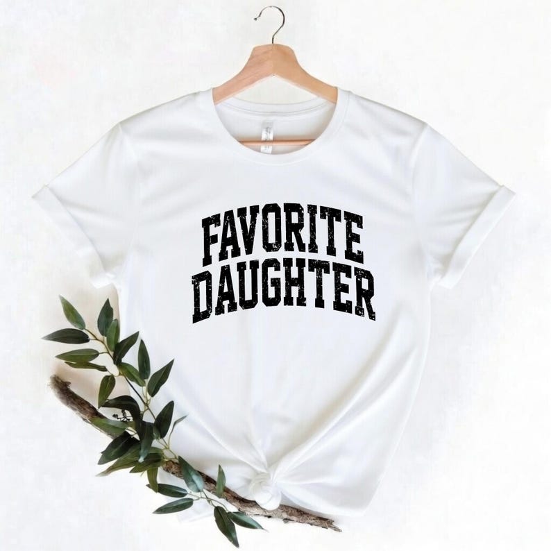 Puede incluir: Una camiseta blanca con el texto "FAVORITE DAUGHTER" en una fuente negra desgastada. La camiseta est&aacute; colgada de una percha de madera. Hay vegetaci&oacute;n en la esquina inferior izquierda de la imagen.