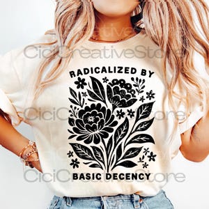 Puede incluir: Camiseta color crema con un diseño floral negro y el texto "RADICALIZED BY BASIC DECENCY". El gráfico incluye flores y hojas estilizadas. La camiseta tiene un corte holgado y es de algodón.