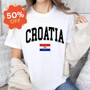 K&ouml;nnte beinhalten: Ein wei&szlig;es Kurzarm-T-Shirt mit dem Wort CROATIA, das in gro&szlig;en schwarzen Gro&szlig;buchstaben in einer geschwungenen Schriftart gedruckt ist, platziert &uuml;ber einer kleinen, rechteckigen kroatischen Flagge mit roten, wei&szlig;en und blauen Streifen und Wappen.