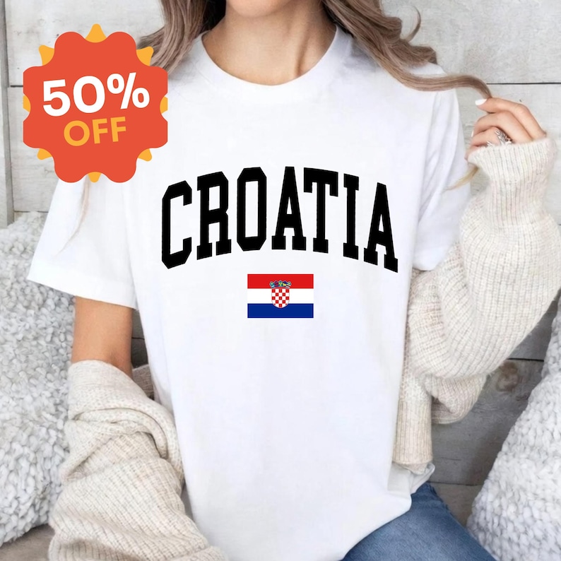 K&ouml;nnte beinhalten: Ein wei&szlig;es Kurzarm-T-Shirt mit dem Wort CROATIA, das in gro&szlig;en schwarzen Gro&szlig;buchstaben in einer geschwungenen Schriftart gedruckt ist, platziert &uuml;ber einer kleinen, rechteckigen kroatischen Flagge mit roten, wei&szlig;en und blauen Streifen und Wappen.