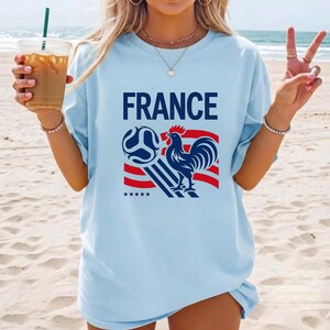 Puede incluir: Una camiseta azul claro con el texto FRANCE en letras azul oscuro sobre un gr&aacute;fico de un gallo y un bal&oacute;n de f&uacute;tbol. El dise&ntilde;o incluye rayas rojas y azules y cinco estrellas peque&ntilde;as debajo del gallo. La camiseta tiene cuello redondo.