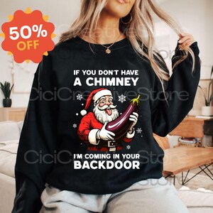 May include: Black sweatshirt with a Santa Claus graphic holding an eggplant. The text reads, "IF YOU DON'T HAVE A CHIMNEY, I'M COMING IN YOUR BACKDOOR." A red and orange 50% OFF sticker is in the upper left corner.