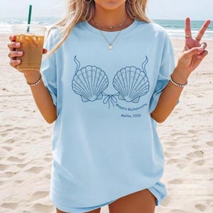 May include: A light blue t-shirt featuring a dark blue line art graphic of two seashells arranged like a bikini top. Below the shells, the text reads "Megan's Bachelorette" and "Malibu, 2026" in a simple, dark blue sans-serif font.
