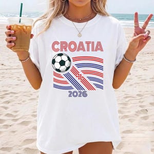 May include: A white short-sleeved t-shirt featuring the text CROATIA in red, a black and white soccer ball, and a graphic design of red and blue horizontal lines forming a flag-like pattern, with the year 2026 printed in blue at the bottom.