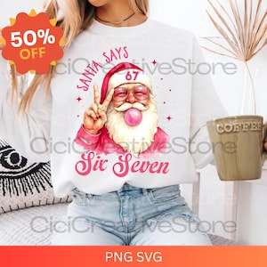 Op de afbeelding: Wit sweatshirt met een afbeelding van de Kerstman met een roze zonnebril en een kauwgomballenbel. De tekst "Santa Says Six Seven" staat in roze letters. Een sticker met 50% korting staat linksboven.