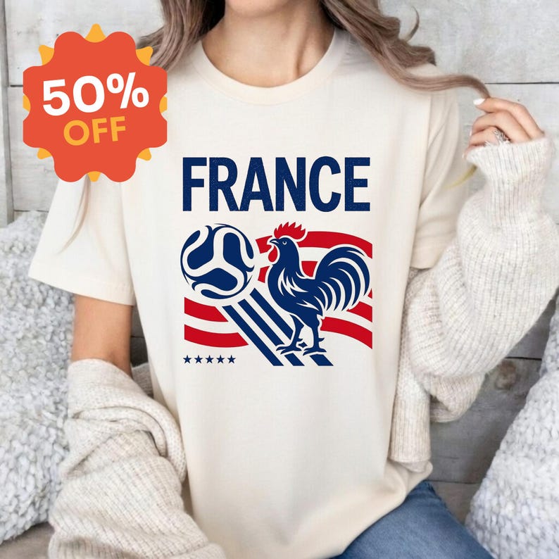 Puede incluir: Una camiseta de manga corta de color crema con un estampado gr&aacute;fico que incluye la palabra FRANCE en azul, un bal&oacute;n de f&uacute;tbol azul, un gallo azul y rayas horizontales rojas. Se ve una insignia naranja en forma de estrella con 50% OFF.