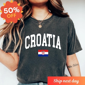 K&ouml;nnte beinhalten: Ein anthrazitfarbenes Kurzarm-T-Shirt mit dem Wort CROATIA in gro&szlig;en, wei&szlig;en Gro&szlig;buchstaben, die bogenf&ouml;rmig &uuml;ber der Brust angeordnet sind, &uuml;ber einer kleinen rechteckigen Grafik der kroatischen Nationalflagge mit roten, wei&szlig;en und blauen Streifen.