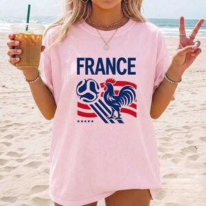 Puede incluir: Una camiseta de manga corta de color rosa claro que presenta la palabra FRANCE en letras azules negritas sobre un gr&aacute;fico de un gallo azul, un bal&oacute;n de f&uacute;tbol azul y rayas rojas y azules con cinco estrellas peque&ntilde;as debajo del dise&ntilde;o.