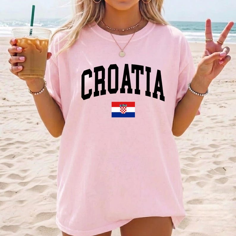 K&ouml;nnte beinhalten: Ein hellrosa T-Shirt mit kurzen &Auml;rmeln, auf dessen Brust das Wort CROATIA in gro&szlig;en schwarzen Gro&szlig;buchstaben gedruckt ist, platziert &uuml;ber einer kleinen rechteckigen Grafik der kroatischen Nationalflagge mit roten, wei&szlig;en und blauen Streifen.
