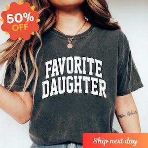 Puede incluir: Camiseta gris oscuro con el texto "FAVORITE DAUGHTER" en letras blancas desgastadas. Una pegatina roja y naranja "50% OFF" est&aacute; en la esquina superior izquierda. La camiseta es de cuello redondo.