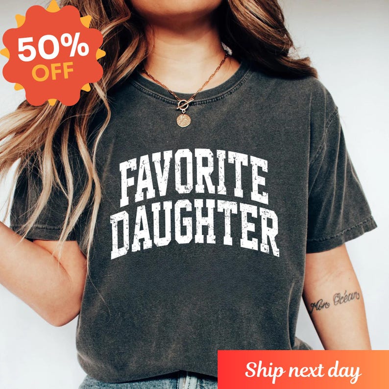 Puede incluir: Camiseta gris oscuro con el texto "FAVORITE DAUGHTER" en letras blancas desgastadas. Una pegatina roja y naranja "50% OFF" est&aacute; en la esquina superior izquierda. La camiseta es de cuello redondo.