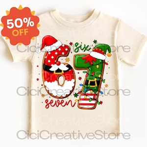 May include: A cream-colored t-shirt featuring a festive design for a child's sixth or seventh birthday. The number '6' is decorated as Santa Claus, and the number '7' is decorated as an elf. The words 'six' and 'seven' are also included.