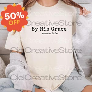 May include: Cream-colored t-shirt with the text "By His Grace" and "romans 3:24" in black. A red and orange 50% OFF sticker is in the upper left corner. The shirt is part of the Creative Store collection.