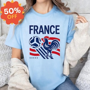 Puede incluir: Una camiseta de manga corta de color azul claro con la palabra FRANCE en letras may&uacute;sculas de color azul oscuro sobre un gr&aacute;fico de un gallo, un bal&oacute;n de f&uacute;tbol y rayas rojas y blancas. Debajo del dise&ntilde;o gr&aacute;fico hay cinco estrellas peque&ntilde;as.