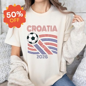 May include: A cream-colored t-shirt featuring a graphic with the text CROATIA and 2026. The design includes a black and white soccer ball and stylized red and blue wavy lines forming a flag-like pattern with a small red and white checkerboard.