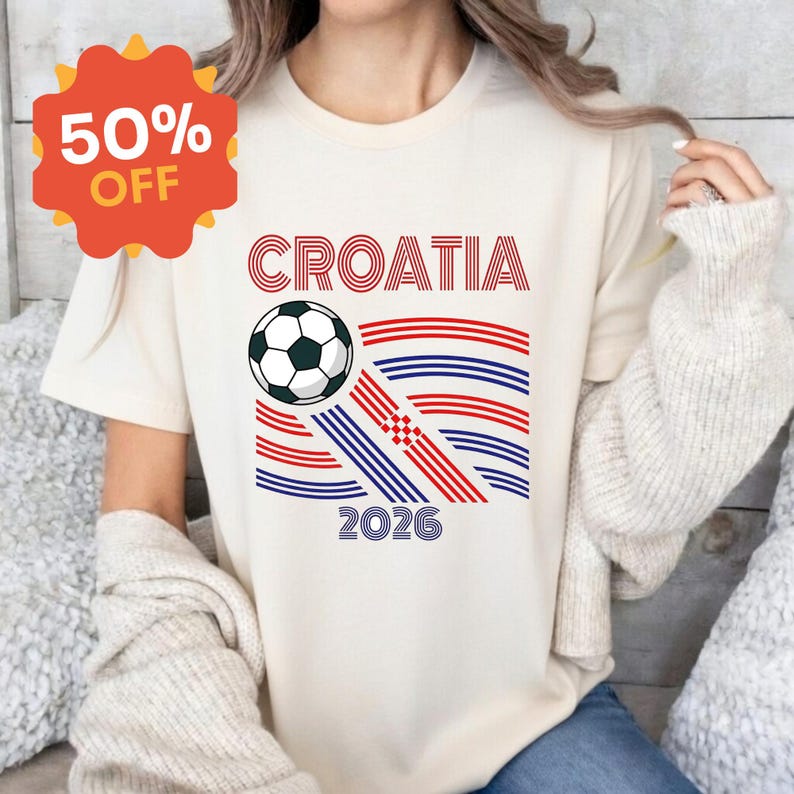 May include: A cream-colored t-shirt featuring a graphic with the text CROATIA and 2026. The design includes a black and white soccer ball and stylized red and blue wavy lines forming a flag-like pattern with a small red and white checkerboard.