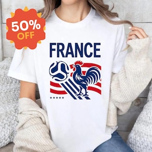 Puede incluir: Una camiseta blanca de manga corta con un estampado gr&aacute;fico que incluye el texto FRANCE en azul, un gallo azul, un bal&oacute;n de f&uacute;tbol y rayas rojas y blancas. Debajo del gallo hay cinco peque&ntilde;as estrellas azules. Una insignia naranja dice 50% OFF.