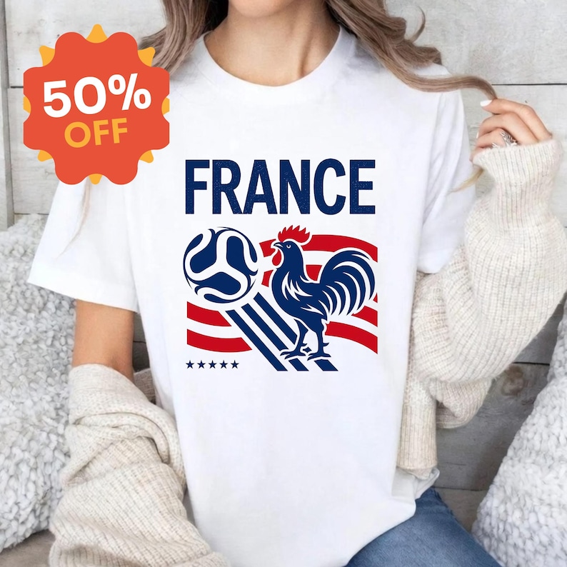 Puede incluir: Una camiseta blanca de manga corta con un estampado gr&aacute;fico que incluye el texto FRANCE en azul, un gallo azul, un bal&oacute;n de f&uacute;tbol y rayas rojas y blancas. Debajo del gallo hay cinco peque&ntilde;as estrellas azules. Una insignia naranja dice 50% OFF.
