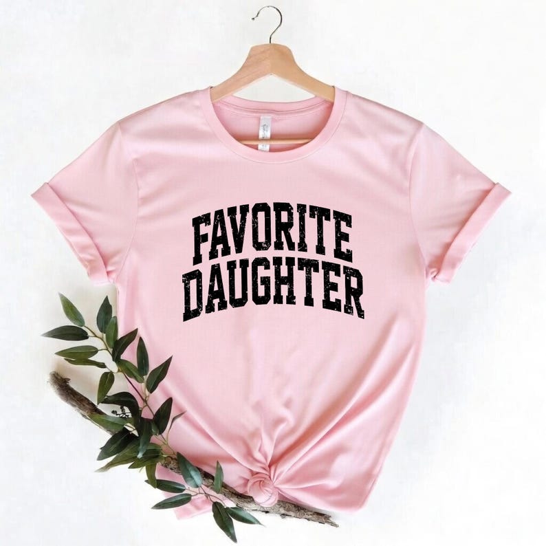 Puede incluir: Camiseta rosa claro con la frase "FAVORITE DAUGHTER" en letras negras y en negrita. La camiseta est&aacute; colgada de una percha de madera. Las mangas est&aacute;n dobladas. El fondo es blanco.
