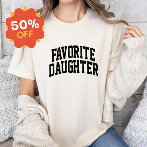 Puede incluir: Camiseta color crema con el texto "FAVORITE DAUGHTER" en letras negras y en negrita. Una pegatina roja y naranja con el 50% de descuento est&aacute; en la esquina superior izquierda. La camiseta se lleva con un su&eacute;ter de color claro.