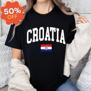 K&ouml;nnte beinhalten: Ein schwarzes Kurzarm-T-Shirt mit dem Wort CROATIA in gro&szlig;en, wei&szlig;en Gro&szlig;buchstaben, die bogenf&ouml;rmig &uuml;ber der Brust angeordnet sind, &uuml;ber einer kleinen rechteckigen Grafik der kroatischen Nationalflagge mit ihren roten, wei&szlig;en und blauen Querstreifen.