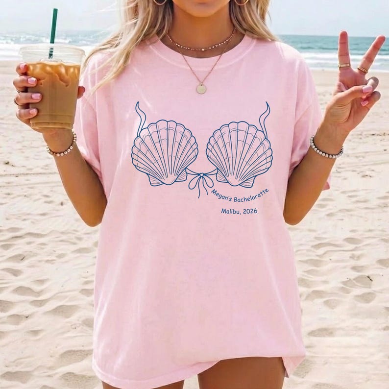 May include: A light pink t-shirt featuring a dark blue graphic of two seashells arranged like a bikini top. Below the shells, the text reads "Megan's Bachelorette" and "Malibu, 2026" in a dark blue font. The shirt has a crew neckline and short sleeves.