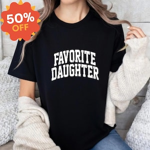 Puede incluir: Camiseta negra con el texto "FAVORITE DAUGHTER" en letras blancas desgastadas. Una pegatina promocional naranja y amarilla con un 50% de DESCUENTO est&aacute; en la esquina superior izquierda. La camiseta la lleva una persona con pelo largo y rubio.