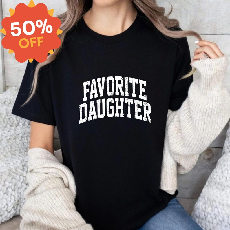 Puede incluir: Camiseta negra con el texto "FAVORITE DAUGHTER" en letras blancas desgastadas. Una pegatina promocional naranja y amarilla con un 50% de DESCUENTO est&aacute; en la esquina superior izquierda. La camiseta la lleva una persona con pelo largo y rubio.