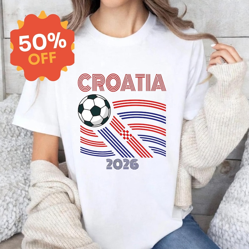 May include: A white short-sleeved t-shirt featuring a graphic print with the text CROATIA and 2026. The design includes a black and white soccer ball and a series of red and blue horizontal lines arranged in a pattern with a small red checkered square.