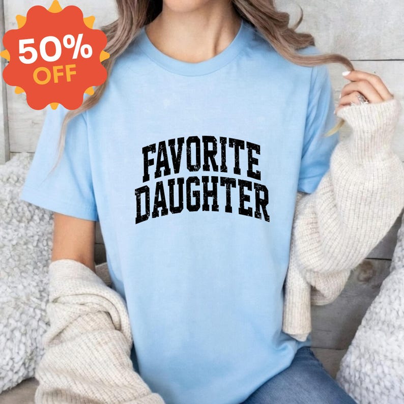 Puede incluir: Camiseta azul claro con la frase "FAVORITE DAUGHTER" en letras negras desgastadas. Una pegatina promocional roja y naranja con el 50% de descuento est&aacute; en la esquina superior izquierda. La camiseta es de manga corta.