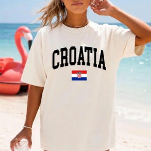 K&ouml;nnte beinhalten: Ein cremefarbenes Kurzarm-T-Shirt mit dem Wort CROATIA in einer fetten, schwarzen, gebogenen Schrift auf der Brust, direkt &uuml;ber einer kleinen, rechteckigen Grafik der kroatischen Nationalflagge mit roten, wei&szlig;en und blauen Streifen.