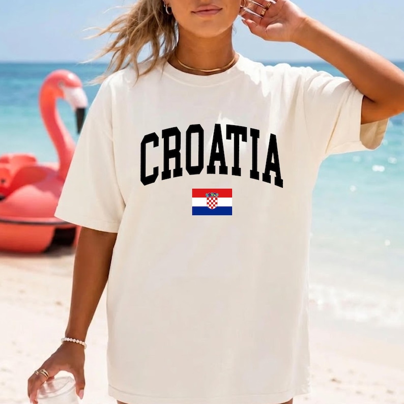 K&ouml;nnte beinhalten: Ein cremefarbenes Kurzarm-T-Shirt mit dem Wort CROATIA in einer fetten, schwarzen, gebogenen Schrift auf der Brust, direkt &uuml;ber einer kleinen, rechteckigen Grafik der kroatischen Nationalflagge mit roten, wei&szlig;en und blauen Streifen.