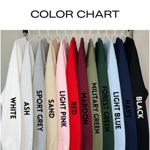 May include: A color chart displaying twelve crewneck sweatshirts hanging on white hangers. The colors are labeled as White, Ash, Sport Grey, Sand, Light Pink, Red, Maroon, Military Green, Forest Green, Light Blue, Navy, and Black in black text.