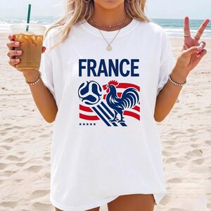 Puede incluir: Una camiseta blanca de manga corta con la palabra FRANCE en letras may&uacute;sculas azules sobre un gr&aacute;fico de un gallo azul, un bal&oacute;n de f&uacute;tbol y rayas rojas y azules. Cinco peque&ntilde;as estrellas azules se sit&uacute;an bajo el dise&ntilde;o en el frente.