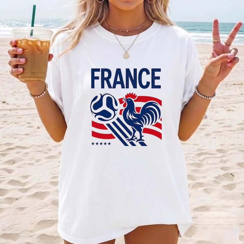 Puede incluir: Una camiseta blanca de manga corta con la palabra FRANCE en letras may&uacute;sculas azules sobre un gr&aacute;fico de un gallo azul, un bal&oacute;n de f&uacute;tbol y rayas rojas y azules. Cinco peque&ntilde;as estrellas azules se sit&uacute;an bajo el dise&ntilde;o en el frente.