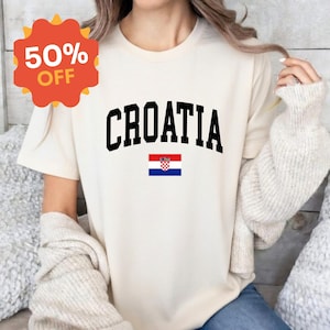 K&ouml;nnte beinhalten: Ein cremefarbenes Kurzarm-T-Shirt mit dem Wort CROATIA in einer fetten, schwarzen, gebogenen Schriftart, platziert &uuml;ber einer kleinen, rechteckigen Grafik der kroatischen Nationalflagge mit ihren horizontalen Streifen in Rot, Wei&szlig; und Blau.