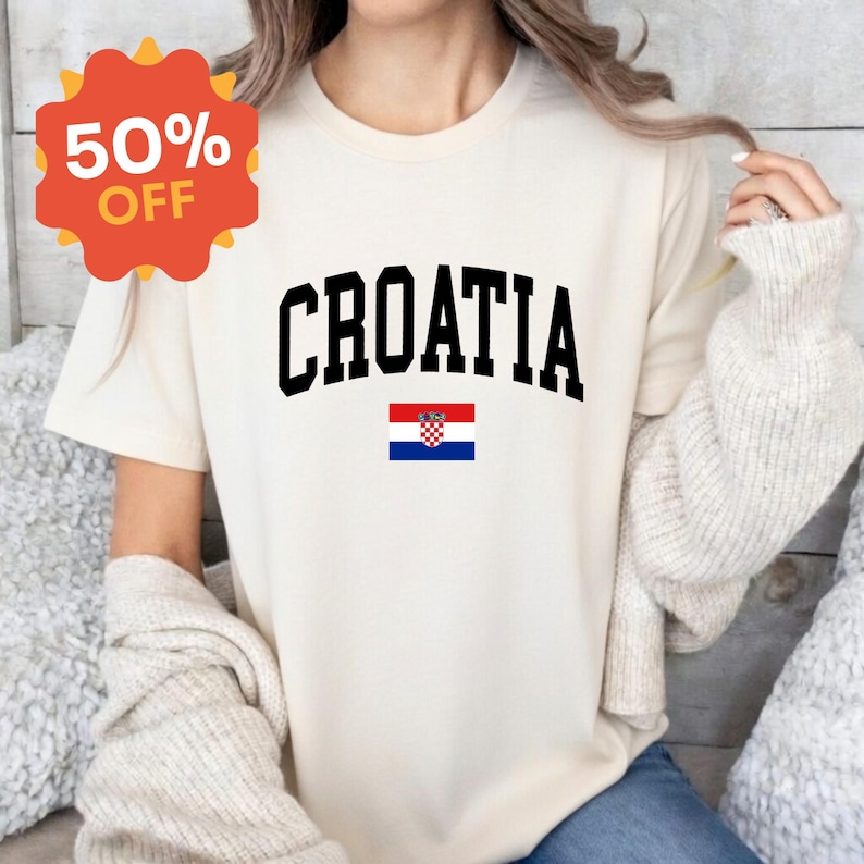 K&ouml;nnte beinhalten: Ein cremefarbenes Kurzarm-T-Shirt mit dem Wort CROATIA in einer fetten, schwarzen, gebogenen Schriftart, platziert &uuml;ber einer kleinen, rechteckigen Grafik der kroatischen Nationalflagge mit ihren horizontalen Streifen in Rot, Wei&szlig; und Blau.