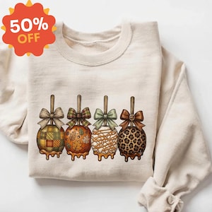 May include: A cream-colored crewneck sweatshirt featuring a graphic of four caramel apples on sticks. Each apple has a unique pattern including plaid, autumn leaves, white drizzle, and leopard print, topped with a decorative fabric bow.